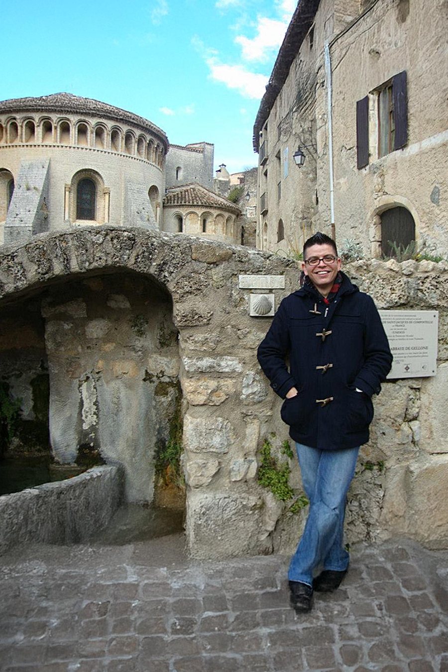 Hector Osuna standing beside a medieval stone bridge and Romanesque abbey in Saint-Guilhem-le-Désert, near Montpellier, France